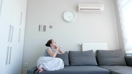 Young brunette woman in her modern apartment in living room. She turns on the air conditioner from the remote control sitting on the sofa. Climate control at home with split system.