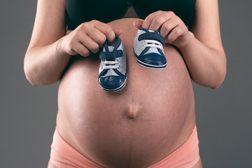 Pregnant woman is holding in hands a baby boy shoes on a gray background. Waiting for the baby to...