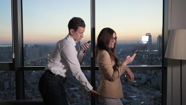 Man touches colleague woman for booty. They are near the panoramic window browsing smartphones during break. She slapped him by the face. Sexual harassment in the office.