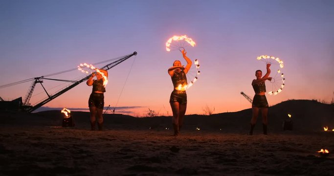 Fire show three women in their hands twist burning spears and fans in the sand with a man with two flamethrowers in slow motion.