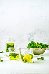 Two glasses with lemon and mint green ice tea, mason jar, and bowl with fresh mint leaves on white background