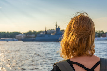 Fototapeta premium Redhead tween tourist girl on Neva river embankment looking at warships and architectural ensemble in summer evening at sunset. Authentic lifestyle moments, travel concept. Saint Petersburg, Russia