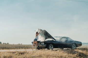 Young female standing near overheated car in the field, bright sunlight, steam under the hood - obrazy, fototapety, plakaty