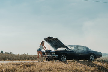Young female standing near overheated car in the field, bright sunlight, steam under the hood