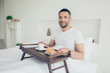 Close up photo amazing he him his macho attractive guy glad early morning sweet bakery breakfast coffee beverage little tea table duvet white sleep wear t-shirt lying bed bright room house indoors