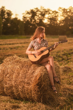 a beautiful, young girl in denim shorts and a plaid shirt plays guitar and lies on a haystack against the sunset sky and a wheat field