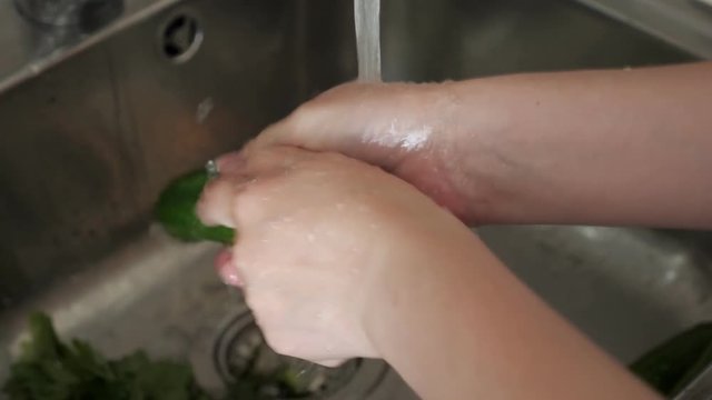 Woman is washing fresh organic vegetables tomatoes and cucumbers for salad under running water in kitchen sink, hands closeup.