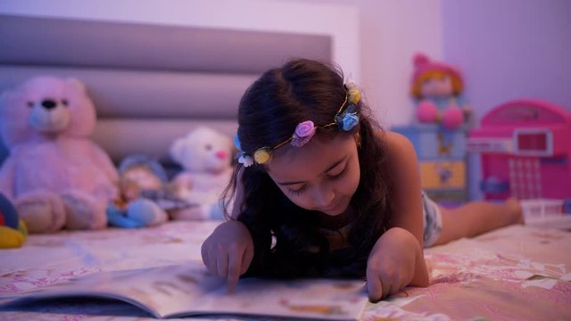 Cute little child girl doing homework and reading a book - Education and smart learning concept. Indian stock footage of a kid enjoying and learning at home. Girl laying on the bed wearing casuals