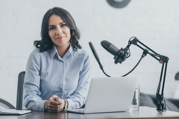beautiful radio host looking at camera while sitting at workplace near microphone and laptop