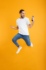 Fototapeta na wymiar Full length photo of joyful caucasian man having beard smiling and holding cellphone while jumping