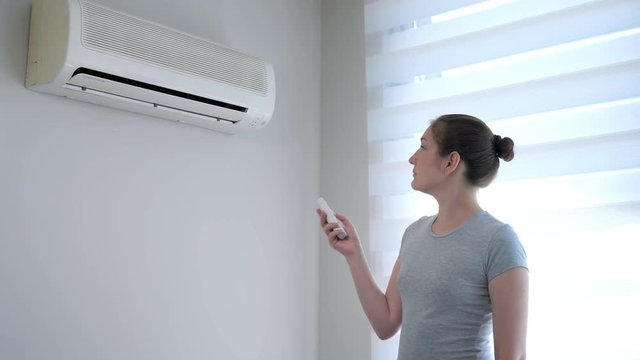 Young brunette woman in her modern apartment. She turns on the air conditioner from the remote control. Climate control at home with split system.
