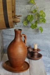 Wooden barrel, earthenware wine jug on plate and candlestick on light wooden table against black concrete wall.