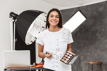 Portrait of happy female makeup artist holding professional cosmetics during fashion photo shooting...