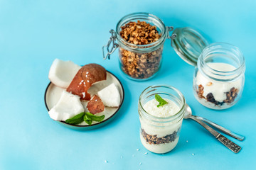 Homemade layered vegan dessert of baked granola and coconut yogurt in glass jar on blue background