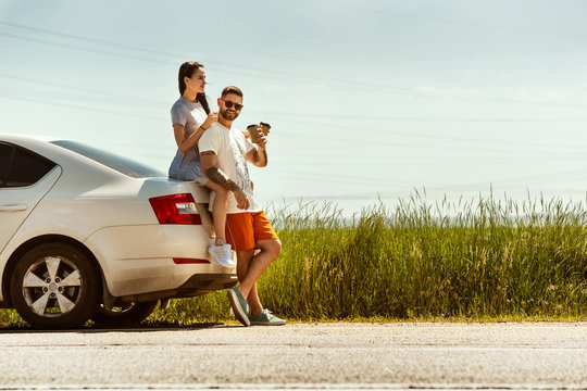 Young couple traveling on the car in sunny summer's day. Woman and man drinking coffee sitting on the trunk, stopping on the way to the sea. Concept of relationship, vacation, summer, holiday, weekend