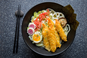 Close up of bowl of gourmet Japanese udon noodle soup with colorful toppings of pickled ginger, boiled egg, swirly pattern narutomaki, and breaded prawns