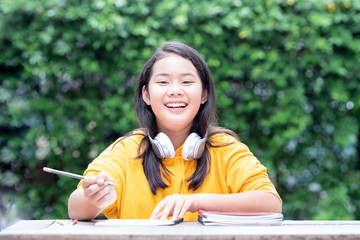 Smiling little Asian schoolgirl doing her homework in the park with headphones on her neck