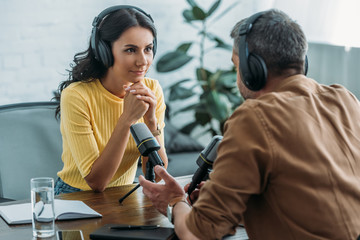 beautiful attentive radio host looking at colleague while sitting at workplace in radio studio