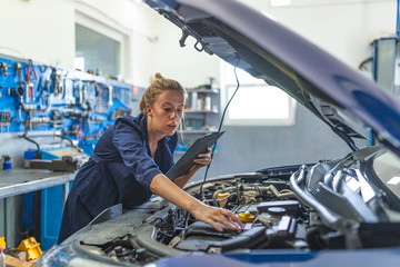 Lovely female auto mechanic smiling, examining engine of an automobile. Cheerful female car technician enjoying working at the garage, copy space. Repair, car service concept