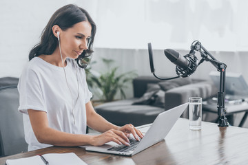 pretty radio host in earphones using laptop while sitting at workplace near microphone