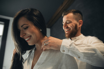 Girl is gathering for a walk. Husband is waiting for wife. Female is preparing for a date. Dressing a newly-married couple. Man wears a beautiful necklace for the woman