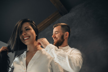 Girl is gathering for a walk. Husband is waiting for wife. Female is preparing for a date. Dressing a newly-married couple. Man wears a beautiful necklace for the woman