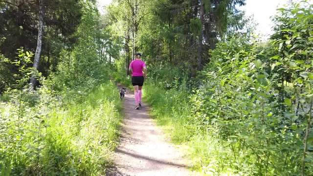 A woman running on a forest trail along the river with her dog on a beautiful sunny summer day. Filmed in realtime at 4k. Camera is following the subject.