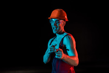 smiling sexy fireman in overall, hardhat and protective goggles pointing with fingers isolated on black