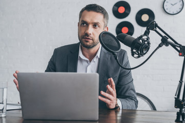 handsome radio host gesturing while speaking in microphone at workplace near laptop