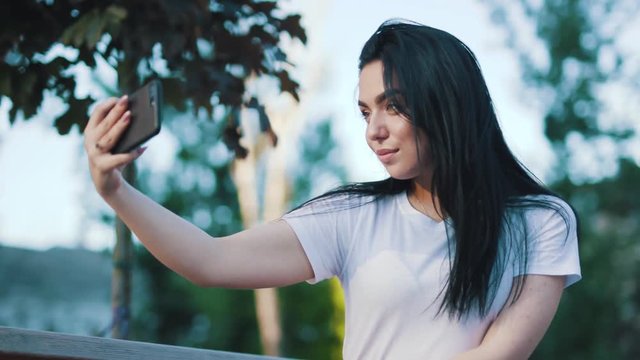 Close up view of beautiful stylish young brunette woman taking selfie via modern phone, posing on camera, making seductive face on nature background. Modern youth, trends concept