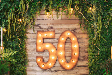 Photo zone made of wood decorated with natural greenery and a garland. The number 50 on a wooden background.