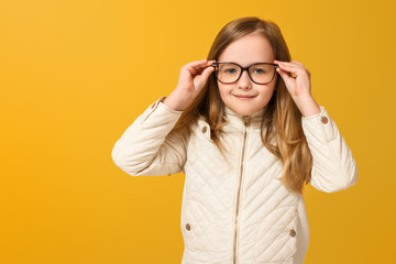 Portrait of a cheerful child in a beige jacket and glasses on a yellow background. Little girl blonde looks into the camera. Autumn concept. Copy space