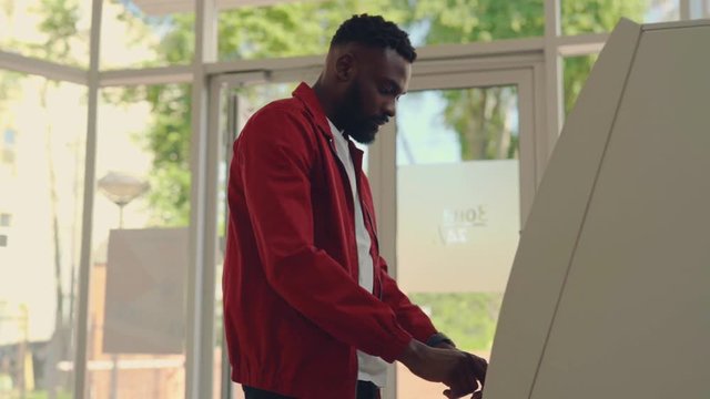 Side view of young stylish Afro-American man inserting credit card in ATM machine, entering code, withdraws cash, machine is broken, money fly out. Shock, problem, stress