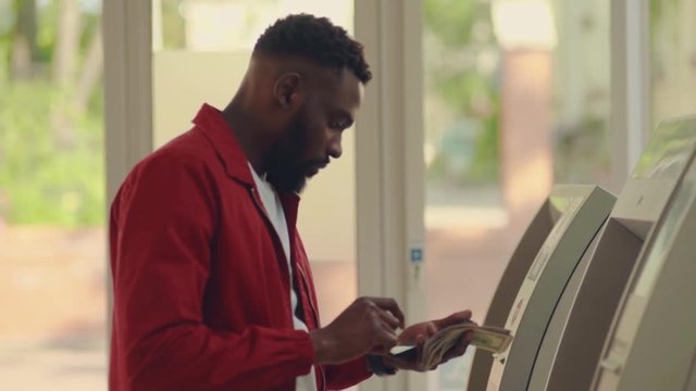 Afro-American man withdraws money from the ATM and checks his balance via phone. Finance, credit card, withdrawal of money. Lifestyle. Grabs money from the ATM