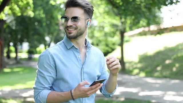 Young happy casual man wearing sunglasses is walking in the park and listens music  on his phone , banging his head as he enjoys it
