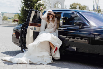 Elegant Bride with Roses Bouquet near Limousine. Sharming Cute Young Woman Dressed in Wedding Garments and Diamond Diadem. Girl Sitting in Black Marriage Car Lift Up Hand with Flower Bunch and Leg