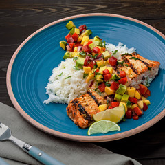 Salmon fillet cooked on grill with exotic sauce made of mango, avocado and pepper on blue plate. Delicious fish meal with jasmine rice on a table.