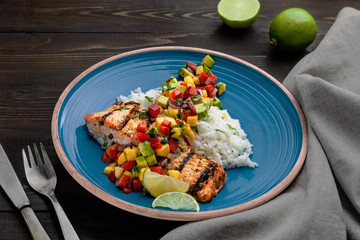 Delicious grilled salmon steak on a blue plate with jasmine rice and sauce made of mango, bell pepper, onion and avocado. Top notch food on a wooden table.