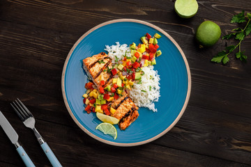 Grilled salmon fillet with fruit sauce made of mango, bell pepper, onion and avocado with basmati rice garnish on a blue plate. Delicious seafood meal. Top view shot above.