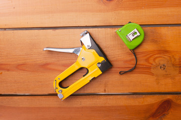 A staple gun and a measuring tape are laying on a wooden surface