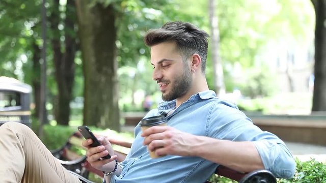 side view of a young casual man sitting on a bench in park and enjoying his morning coffee while reading on news on his smartphone
