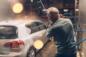 Senior man washing his car in the evening at car wash station using high pressure water.