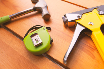 A hammer, measuring tape, and a staple gun are laying on a wooden surface