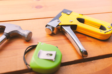 A hammer, measuring tape, and a staple gun are laying on a wooden surface
