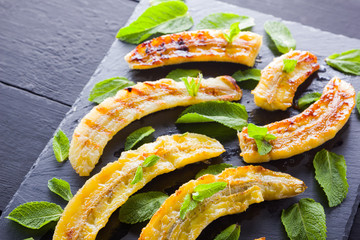 Halves of grilled bananas on a black kitchen board and shale stone. Baked banana dessert with mint...