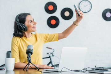 attractive radio host in headphones taking selfie with smartphone while sitting near microphone