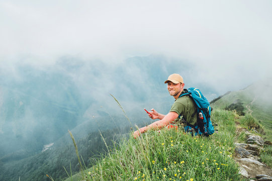 Tired backpacker man browsing internet using smartphone enjoying cloudy valley bottom walking by the foggy cloudy weather mountain range . Active sports trekking and navigation applications concept.
