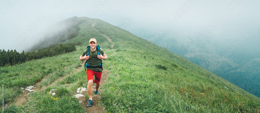 Wall mural smiling hiker man walking by the foggy cloudy weather mountain range path with backpack. active spor