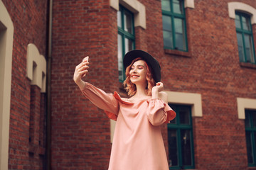 Girl in a beautiful pink dress and black hat. The girl with pink curly hair travels. A girl makes a photo on a smartphone. Girl making selfie on tour in Europe.