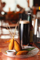 Corn dogs on sticks with chicken sausage, deep fried in egg batter, served with balsamic vinegar and a glass of cold stout, on bokeh background